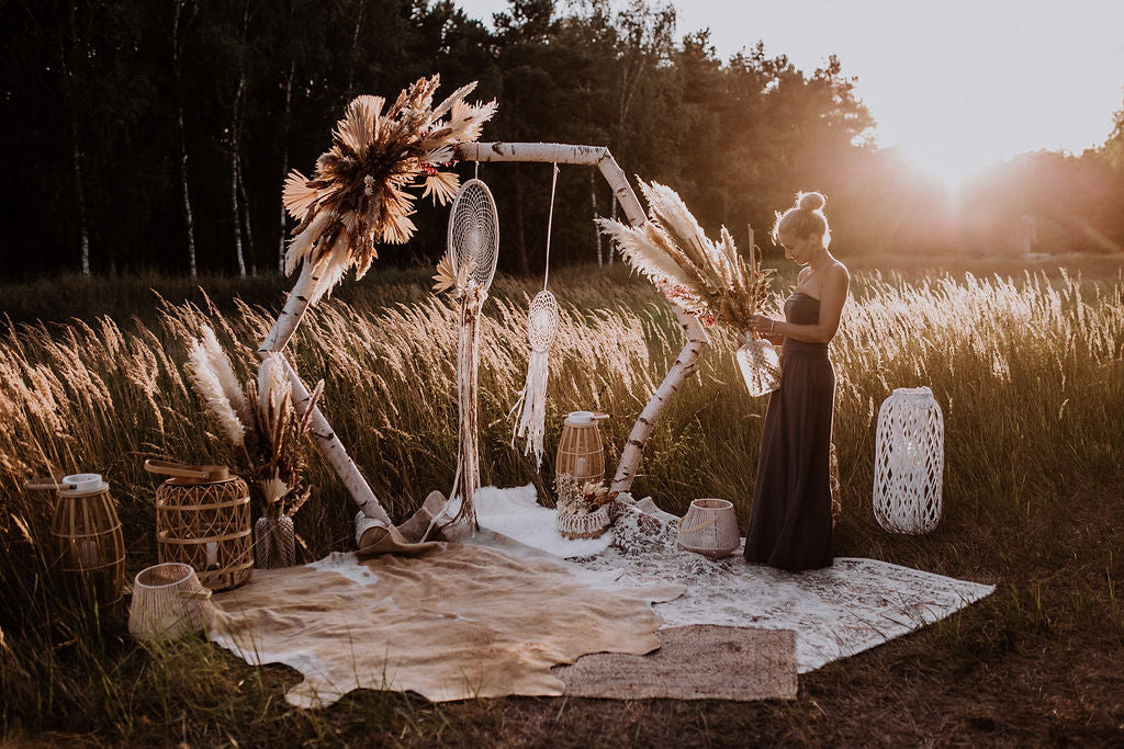 Sabrina Buzwan von Blütenmomente dekoriert einen Traubogen aus Birkenzweigen auf einem Feld mit Trockenblumen im Boho-Stil